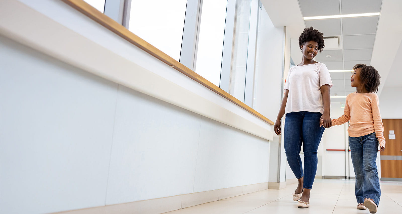 A smiling mother and daughter walking the corridor of a hospital holding hands