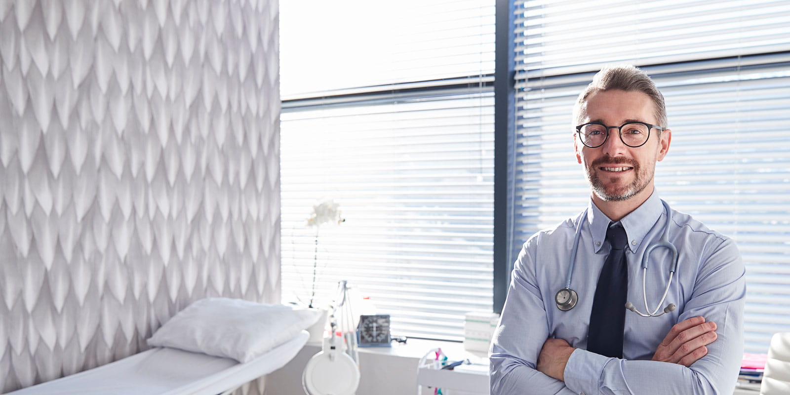 Portrait Of Smiling Mature Male Doctor With Stethoscope Standing By Desk In Office