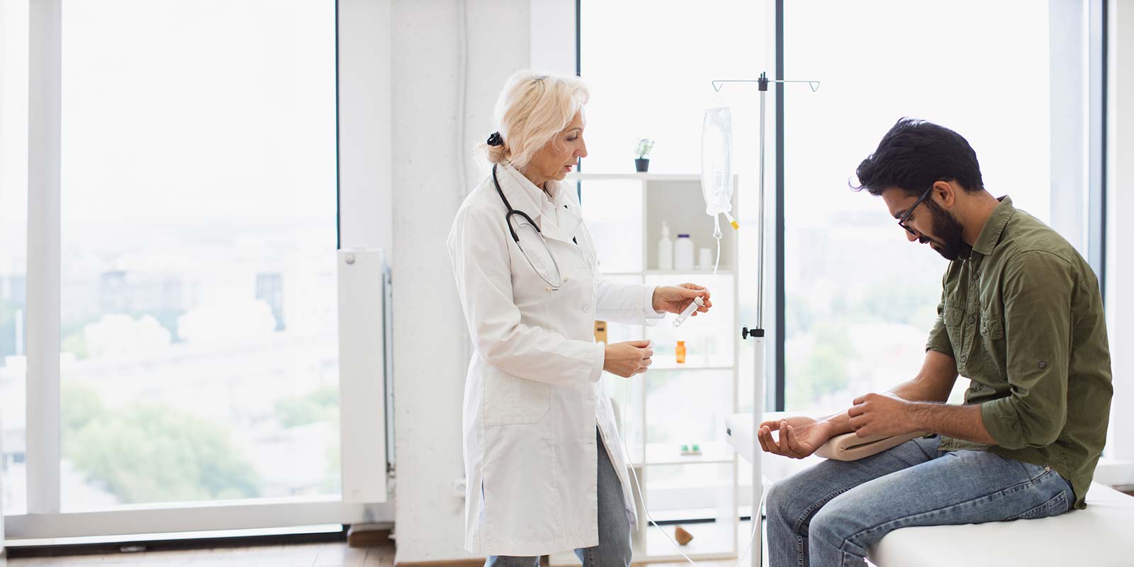 Doctor checking solution for infusion of male patient sitting on exam bed