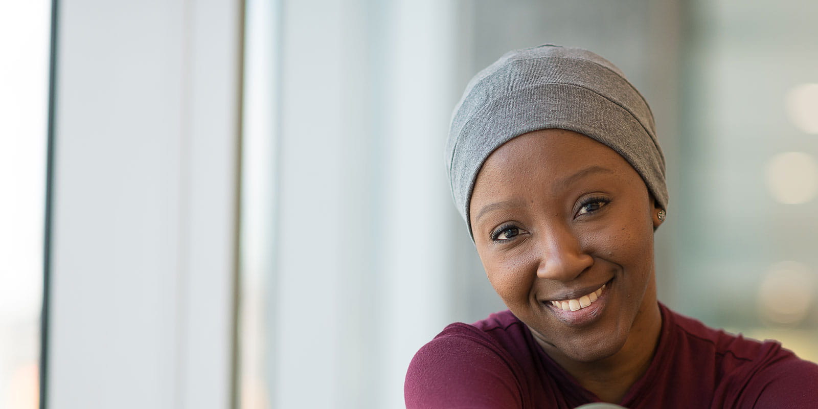 A smiling woman with cancer wearing a hat is seated near a large bay of windows