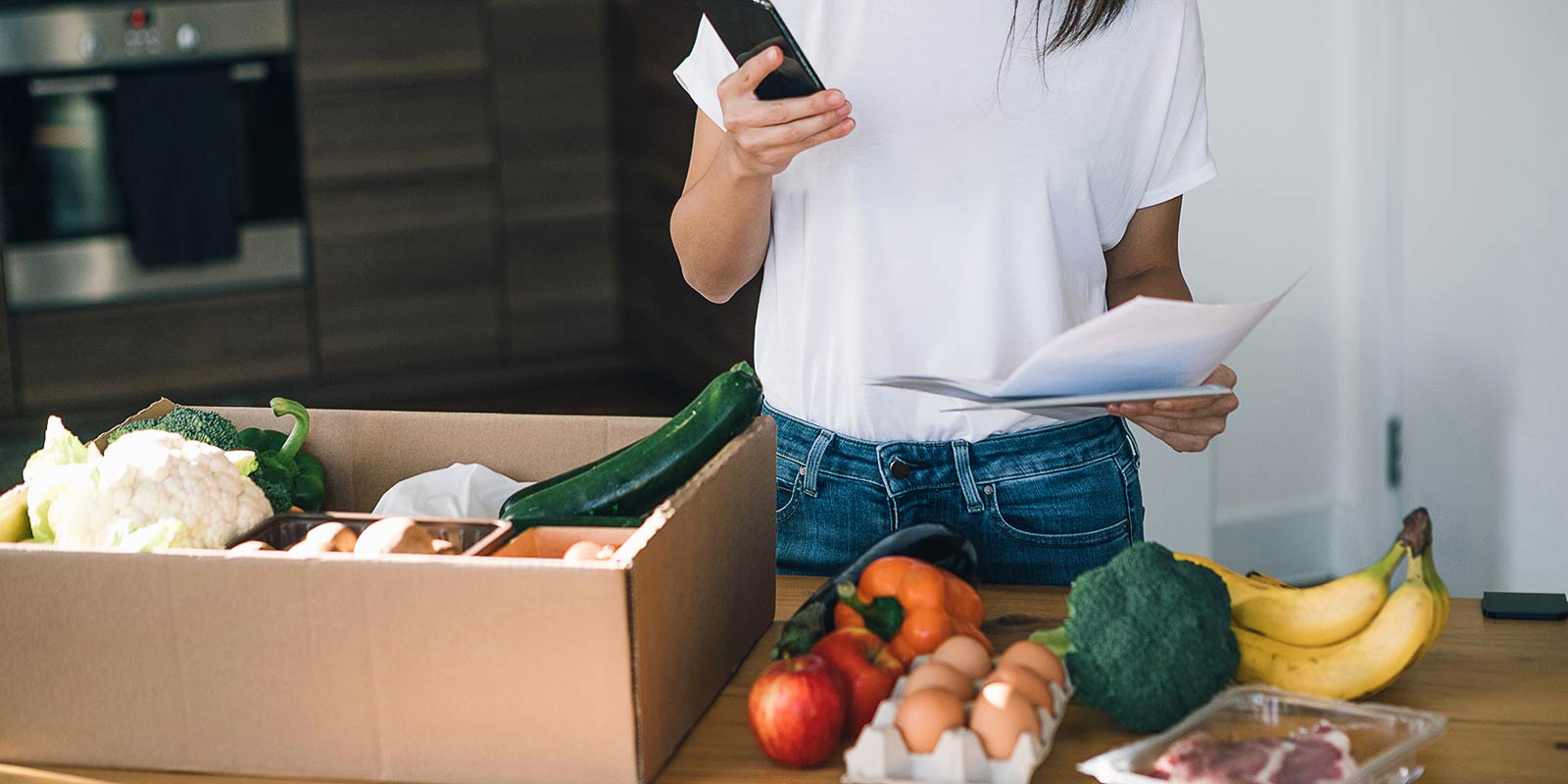 oung woman checking delivery receipt with smartphone while laying out fresh organic fruits, vegetables, meat and dairy products on dining table from a delivery box