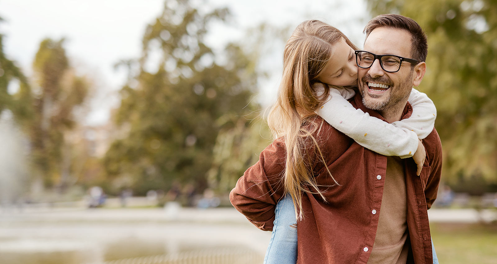 A cute little daughter kissing her happy father while he is giving her a piggyback ride outside