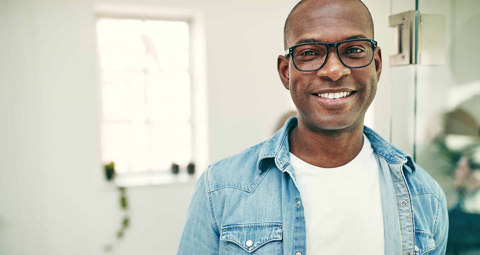Young African businessman wearing glasses and smiling while leaning against a glass wall in a modern office