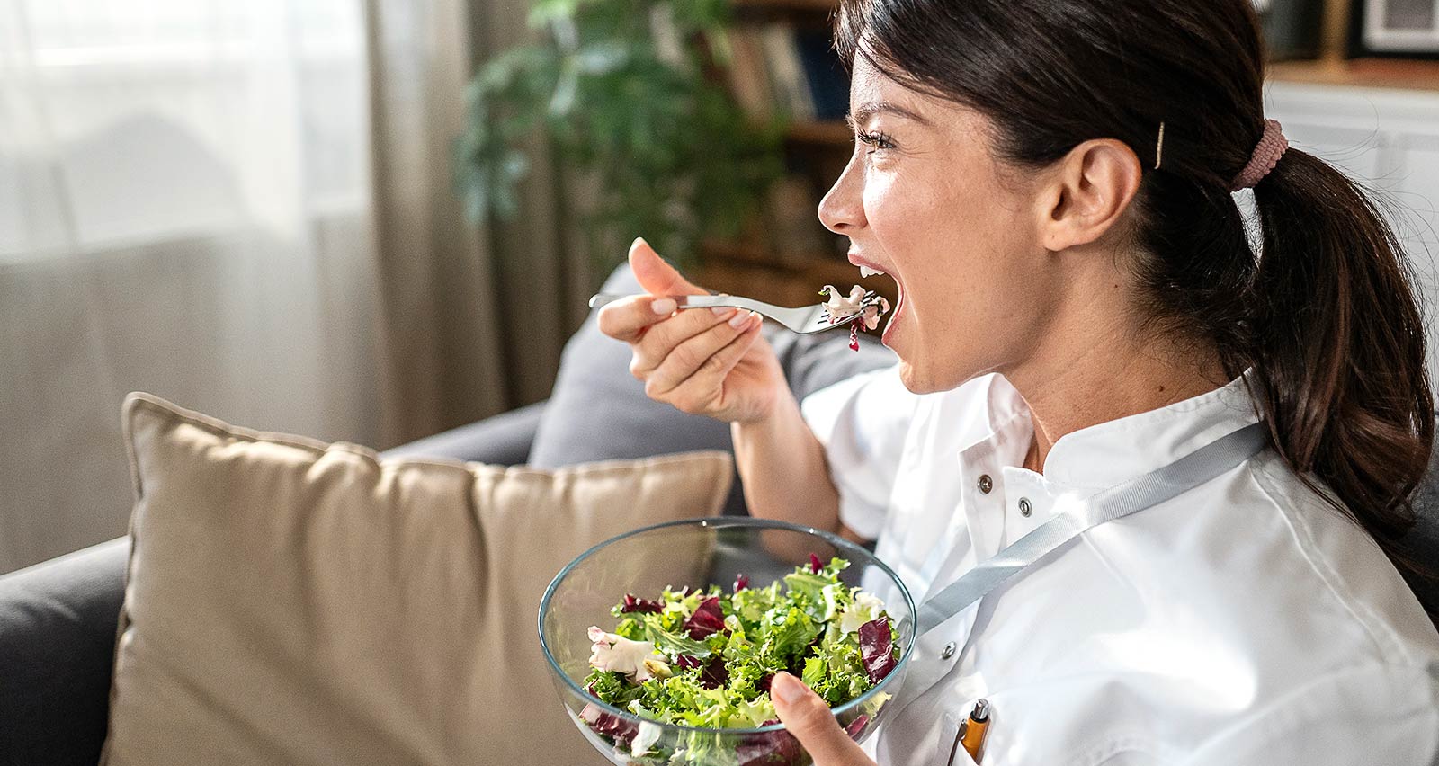 A female healthcare worker in uniform relaxing on a sofa at home, enjoying a fresh salad from a glass bowl during a mindful lunch break