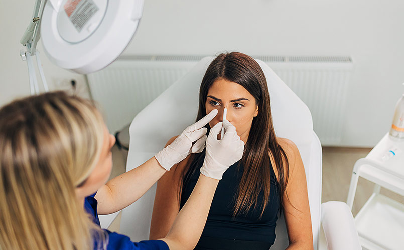 A plastic surgeon examines her young female patient while sketching lines in preparation for a nose job 