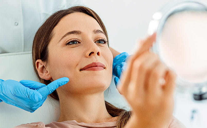 A smiling woman is having a facial treatment while looking at herself in a handheld mirror