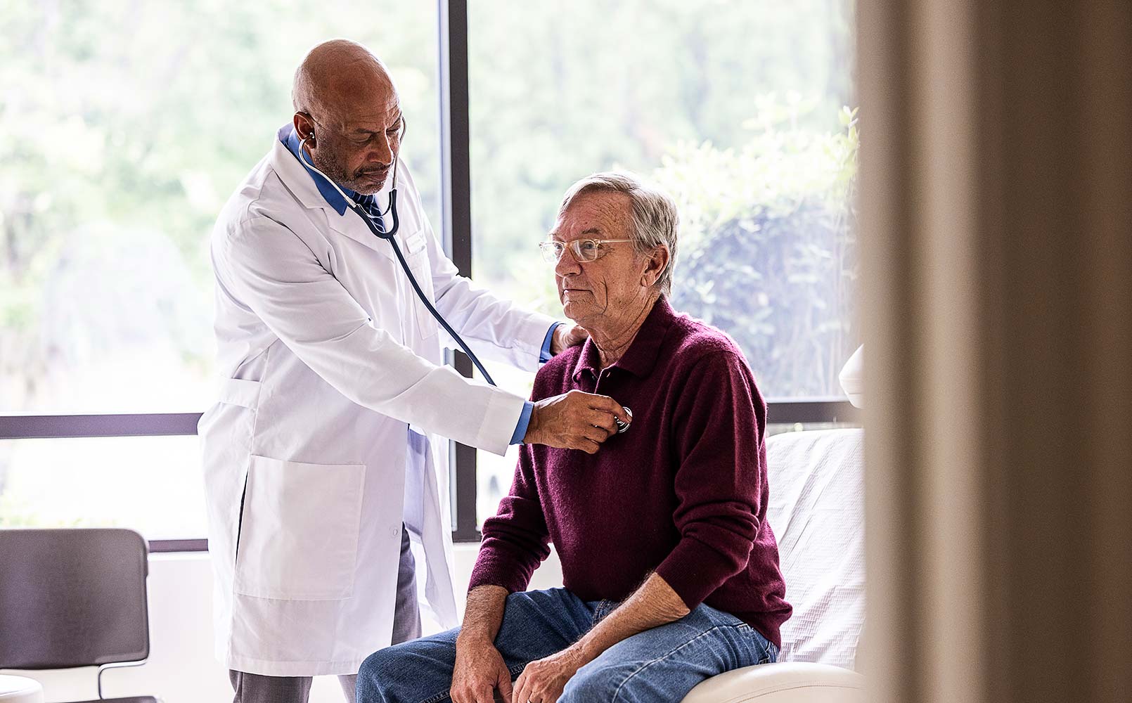 A male doctor listening to senior man’s heart in an exam room