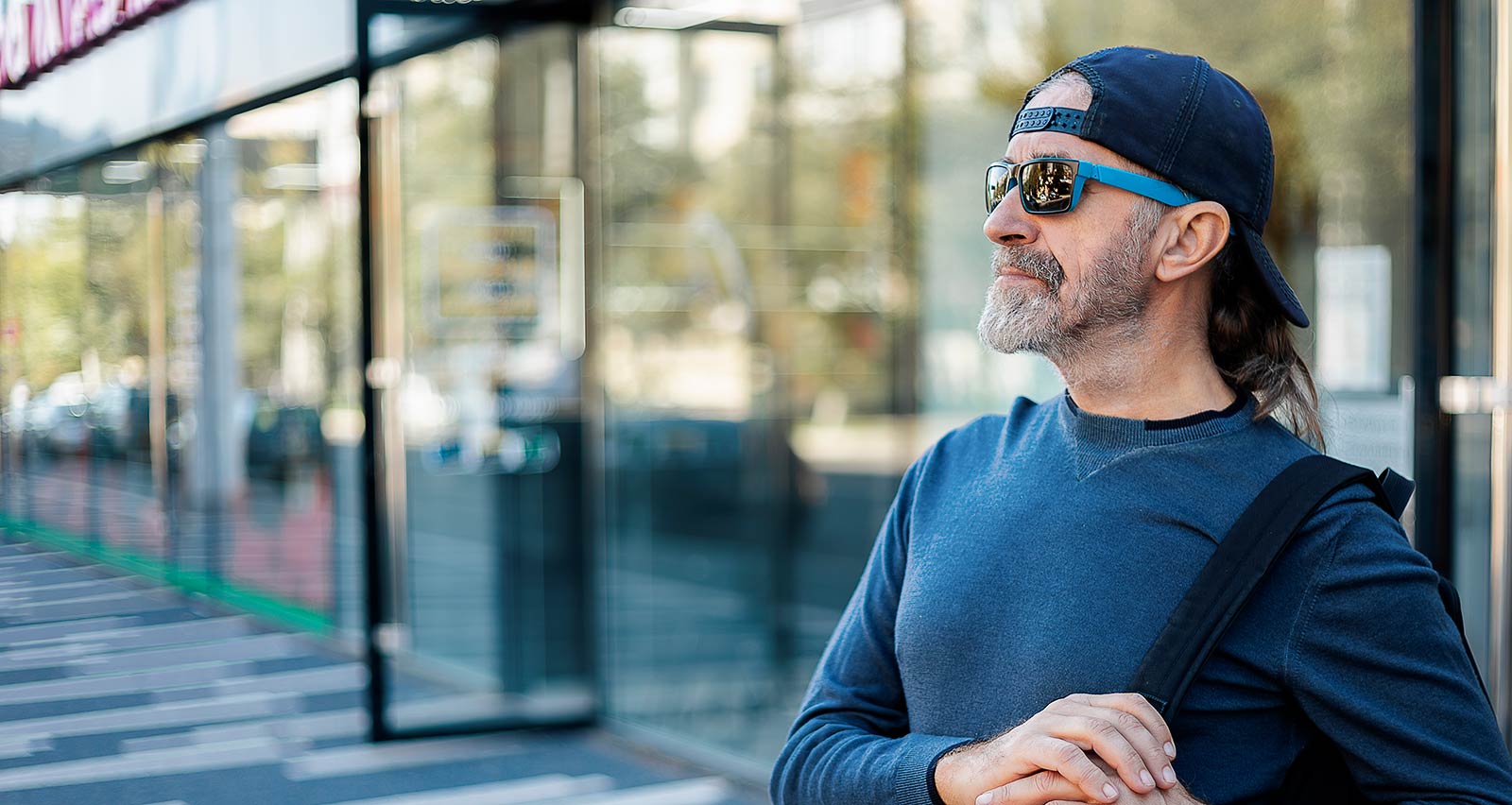 A middle-aged man wearing sunglasses and a cap walks through a city