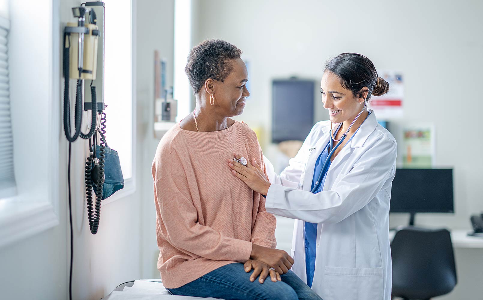 A senior woman of African decent sits up on an exam table as her female doctor listens to her heart during a routine check-up