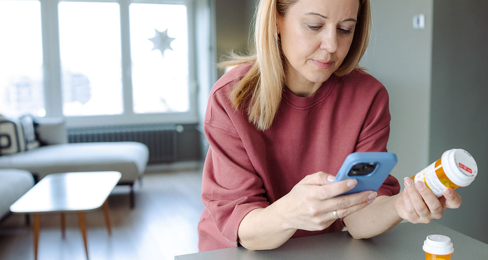 A woman holds a smartphone and a pill bottle while seated in a contemporary apartment