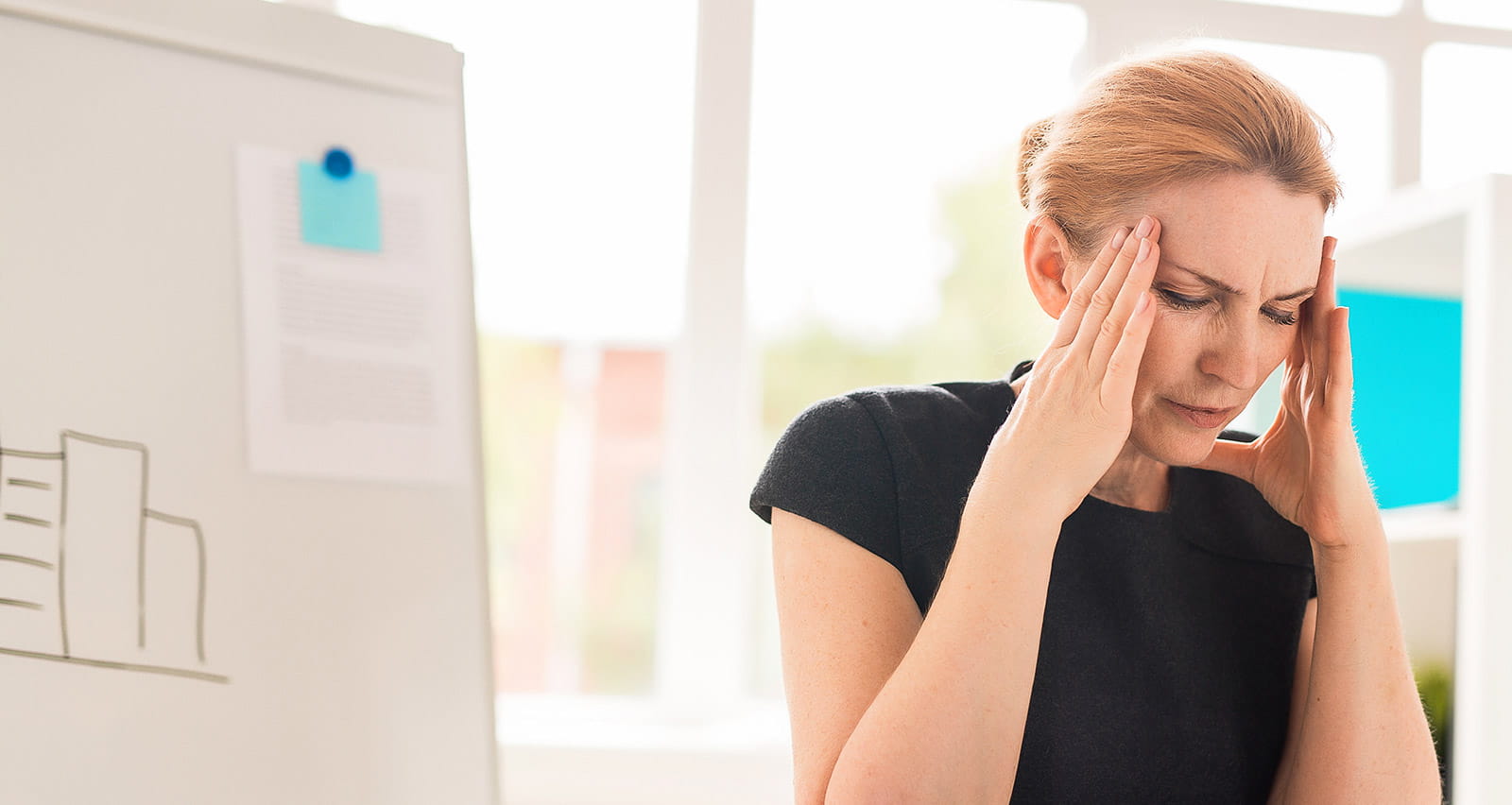 Exhausted middle-aged businesswoman suffering from terrible headache while sitting at desk in spacious office with panoramic windows