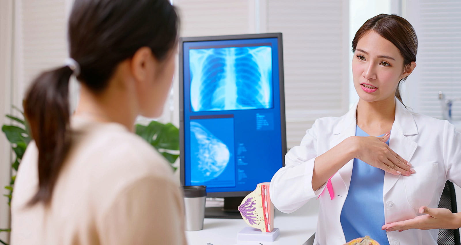 A female doctor wearing a white coat with a pink ribbon explains breast self examination to her patient