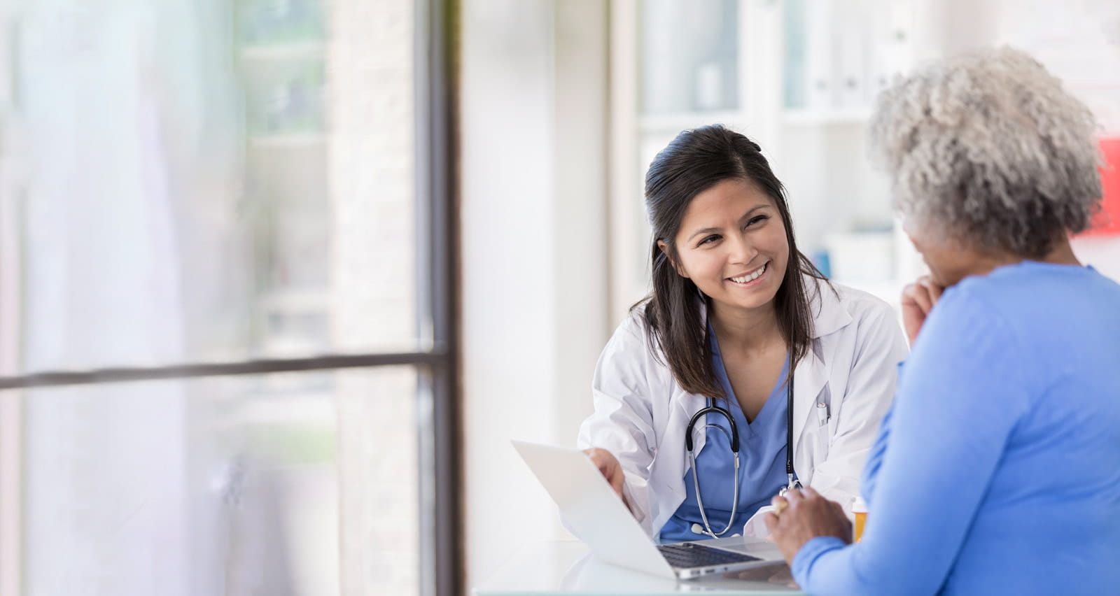 Young nurse uses laptop while sitting with senior patient explaining results
