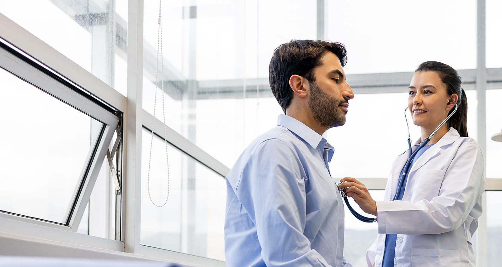 A female doctor examinies a patient at her office and listens to his breathing with a stethoscope