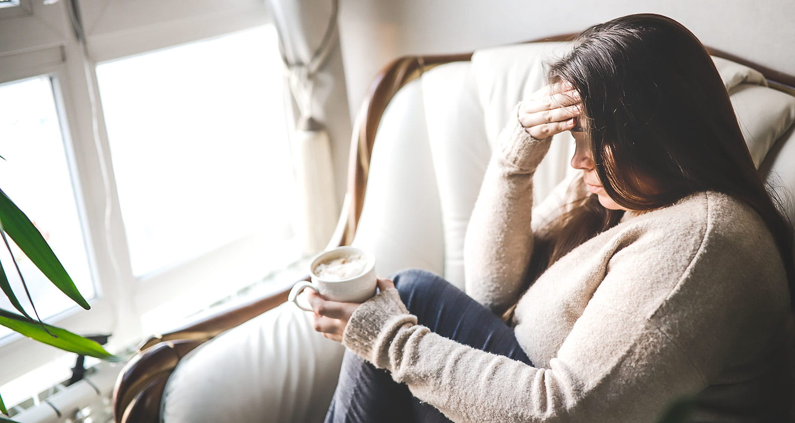 A woman wearing a sweater holds a cup of coffee while gazing out the window holding her head