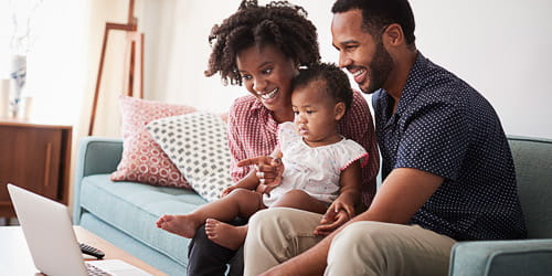 Family With Baby Daughter Sitting On Sofa At Home Looking At Laptop Computer